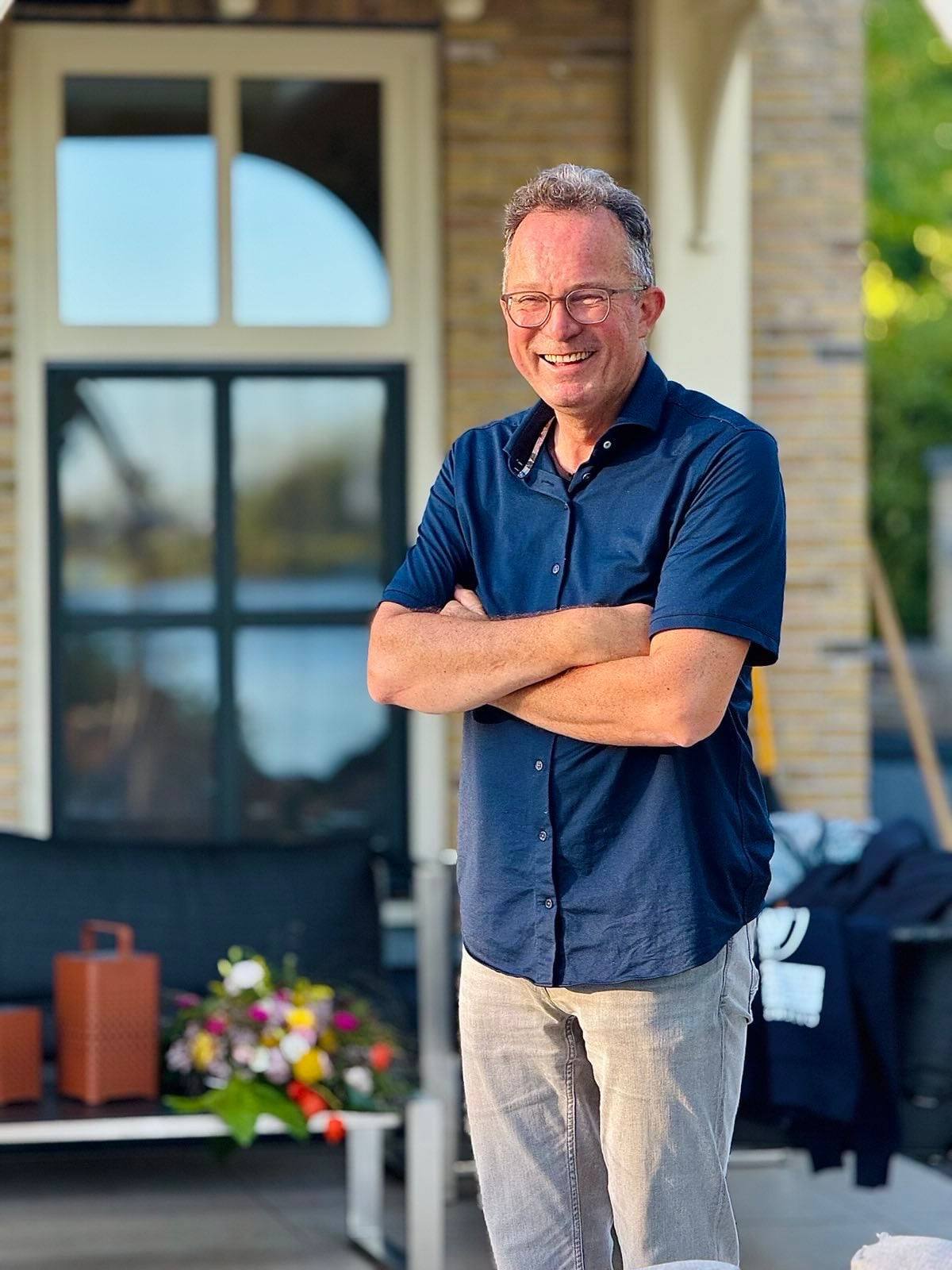 A man in a dark shirt, arms folded, laughing, outside a brick house in evening sun.